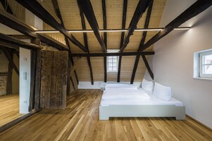 Un dormitorio con techo inclinado, vigas de madera a la vista, un ventanal grande y una cama blanca.