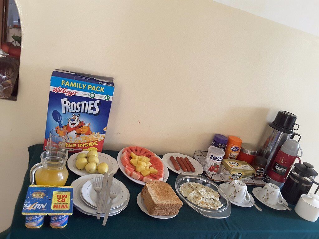 A breakfast table with a Kellogg's Frosties cereal box, plates of fruit, hot dogs, toast, and a coffee pot.
