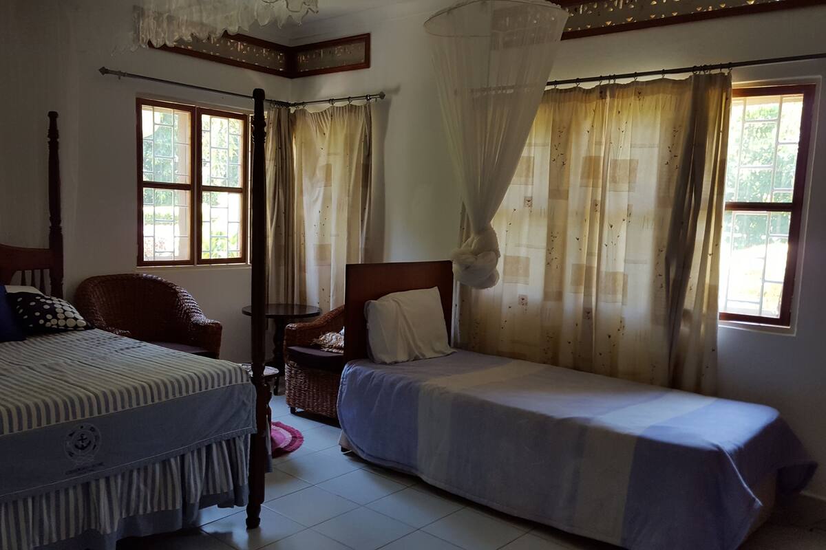 Una habitación con dos camas individuales, una silla de madera, una mesita y una ventana con cortinas.