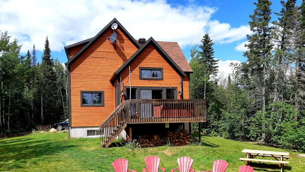 Ein zweistöckiges Holzhaus mit einer Terrasse, umgeben von einem Rasen und Bäumen.