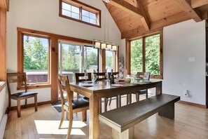 A dining area with a wooden table, chairs, and a bench. Large windows provide natural light and views of greenery outside.