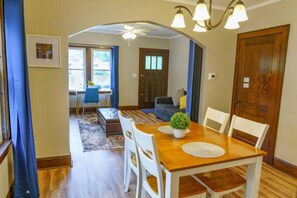 Un coin repas avec une table en bois, des chaises blanches et une petite plante en pot. Un ventilateur de plafond est installé au-dessus de la table, et une porte se trouve sur le côté droit.
