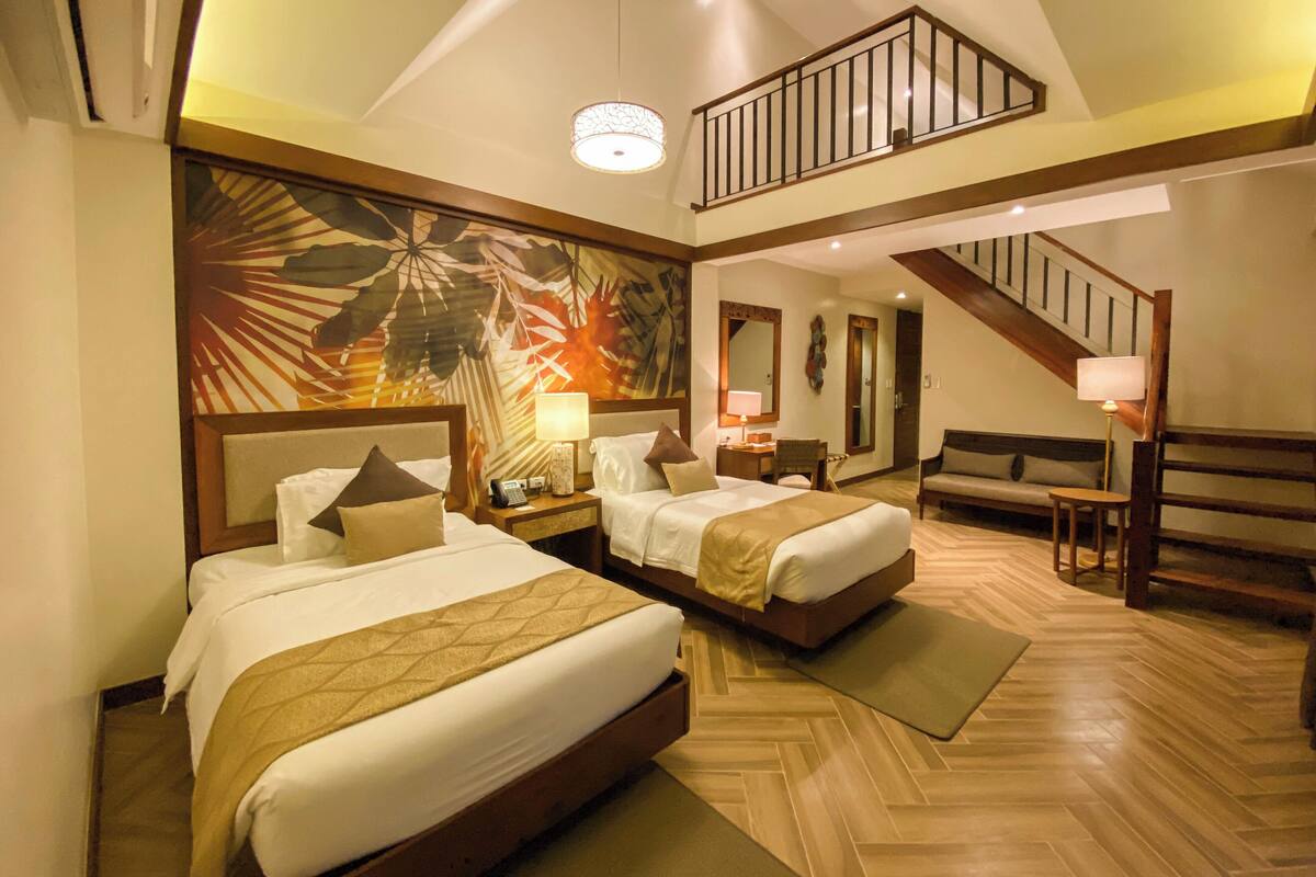 A spacious hotel room with two beds, a large mural, a wooden staircase, and a seating area.
