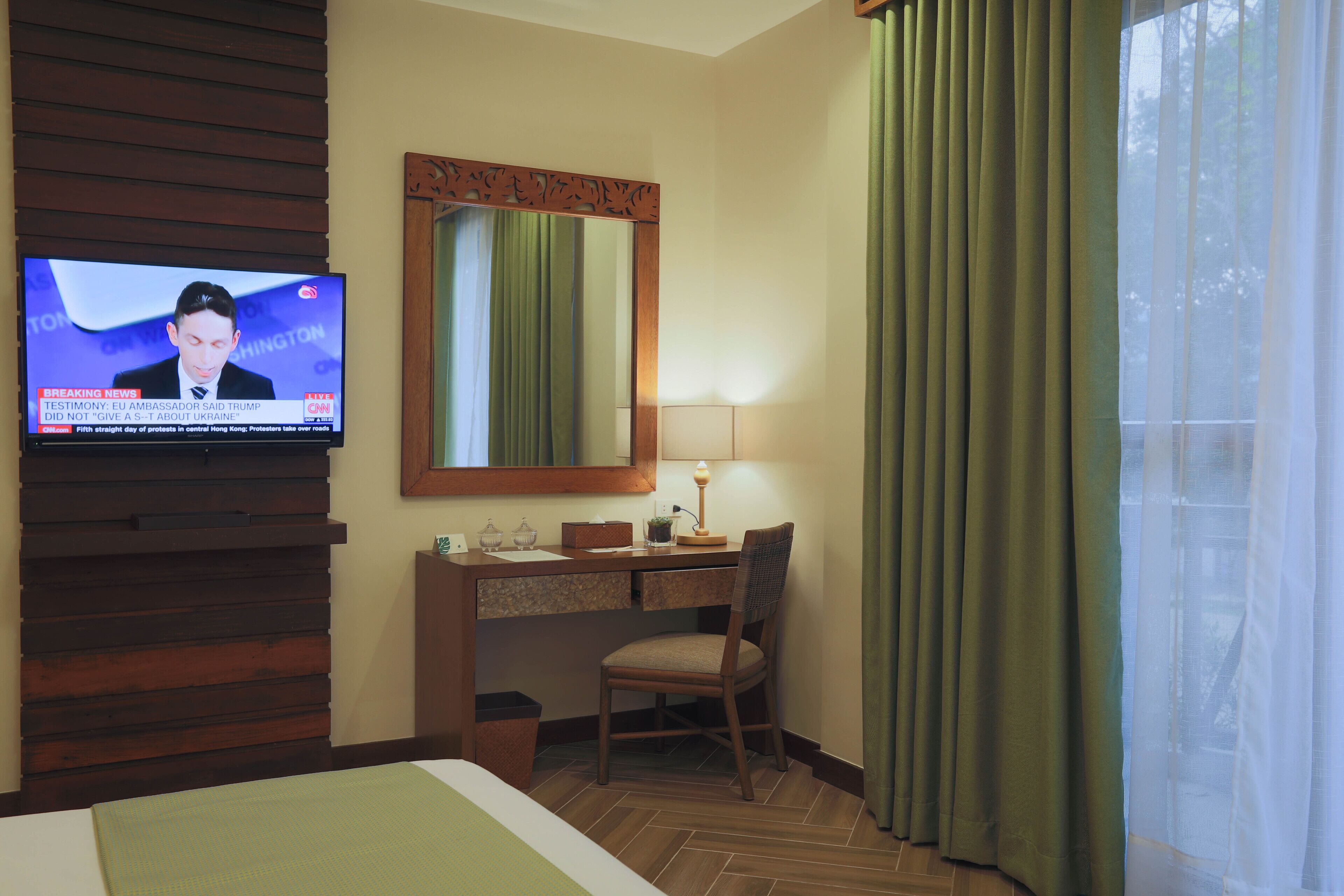 Habitación de hotel con cama, escritorio con silla, televisor montado en la pared, espejo y ventana con cortinas.
