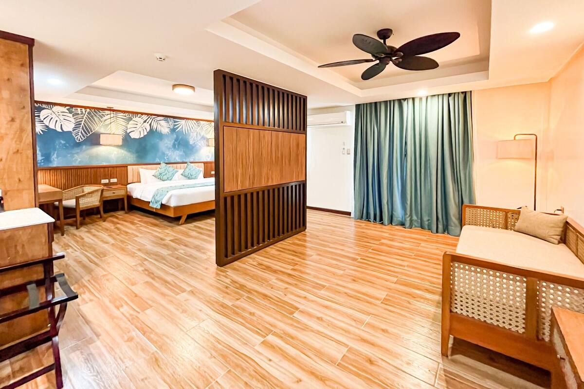 A hotel room with a wooden floor, a bed with a headboard, a desk, a chair, a sofa, a ceiling fan, and a wall with a tropical mural.