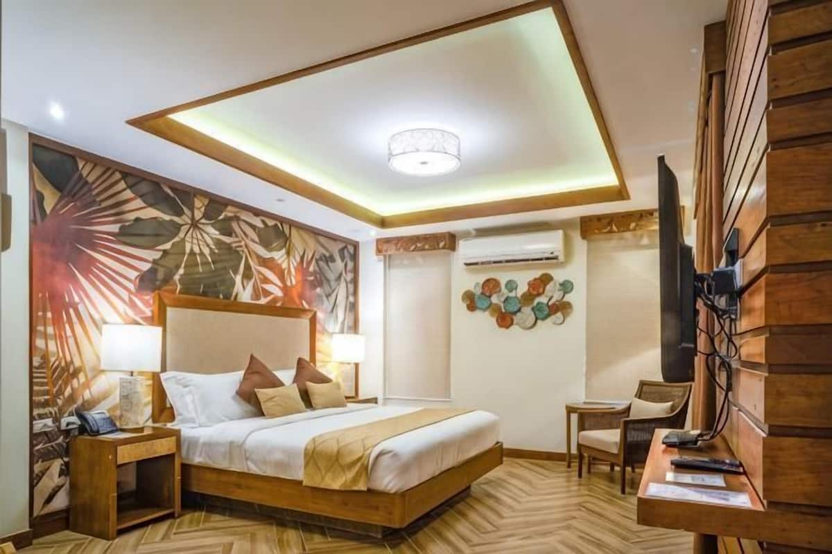 A hotel room with a bed, bedside table, lamp, wall art, television, and a small table with remote controls.