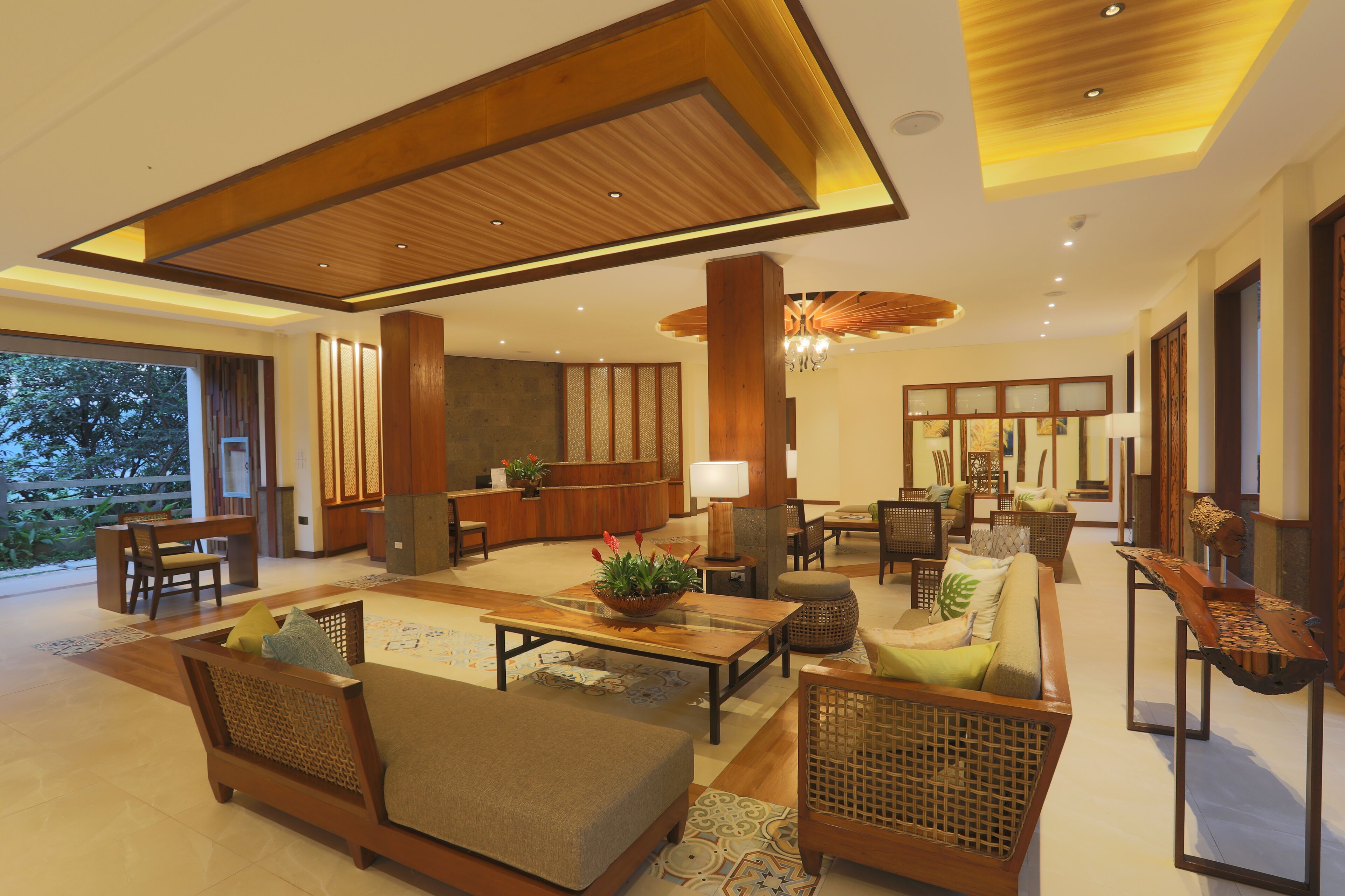 A spacious living area with a wooden ceiling, a reception desk, and a large window offering a view of greenery.