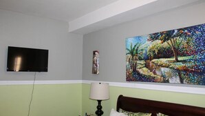 A hotel room with a bed, a TV mounted on the wall, and a painting above the bed. - The Artist Place @ Kensington New Kingston (New Kingston)