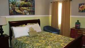 A bedroom with a bed, a chair, a nightstand, a dresser, and a painting on the wall. - The Artist Place @ Kensington New Kingston (New Kingston)