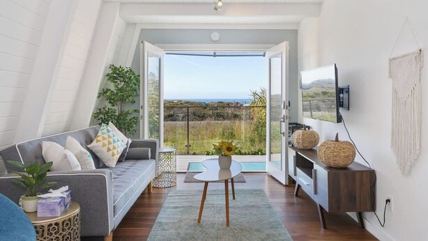 Un salon moderne avec un canapé gris, une table basse et un téléviseur fixé au mur. Une grande fenêtre offre une vue sur la campagne.