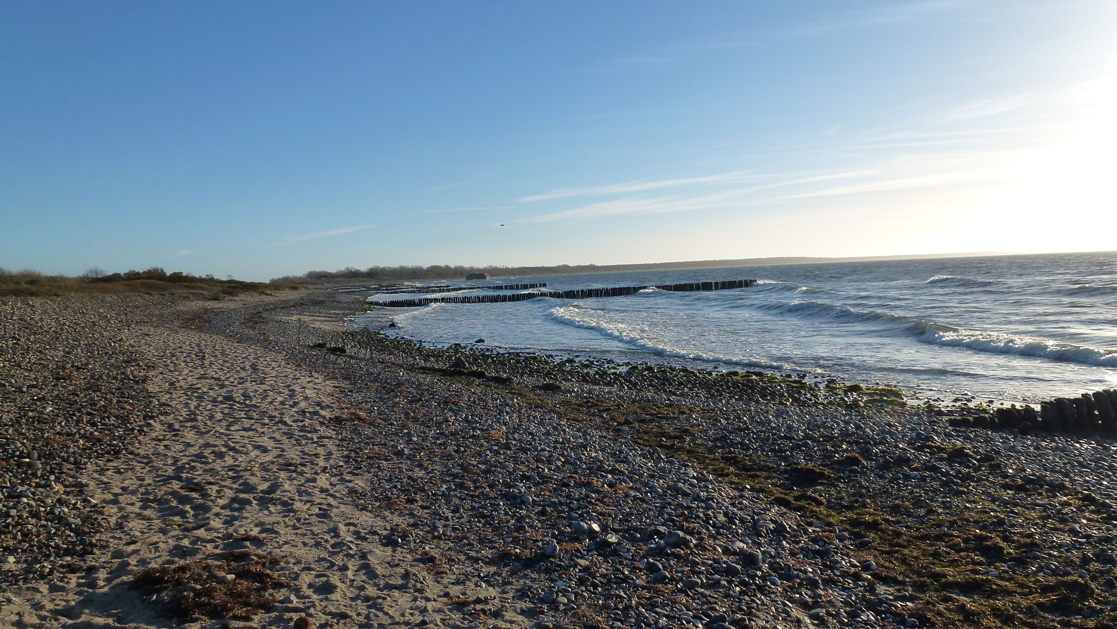 Una spiaggia con ciottoli, con le onde che si infrangono sulla riva e un molo di legno che si protende nell'acqua.