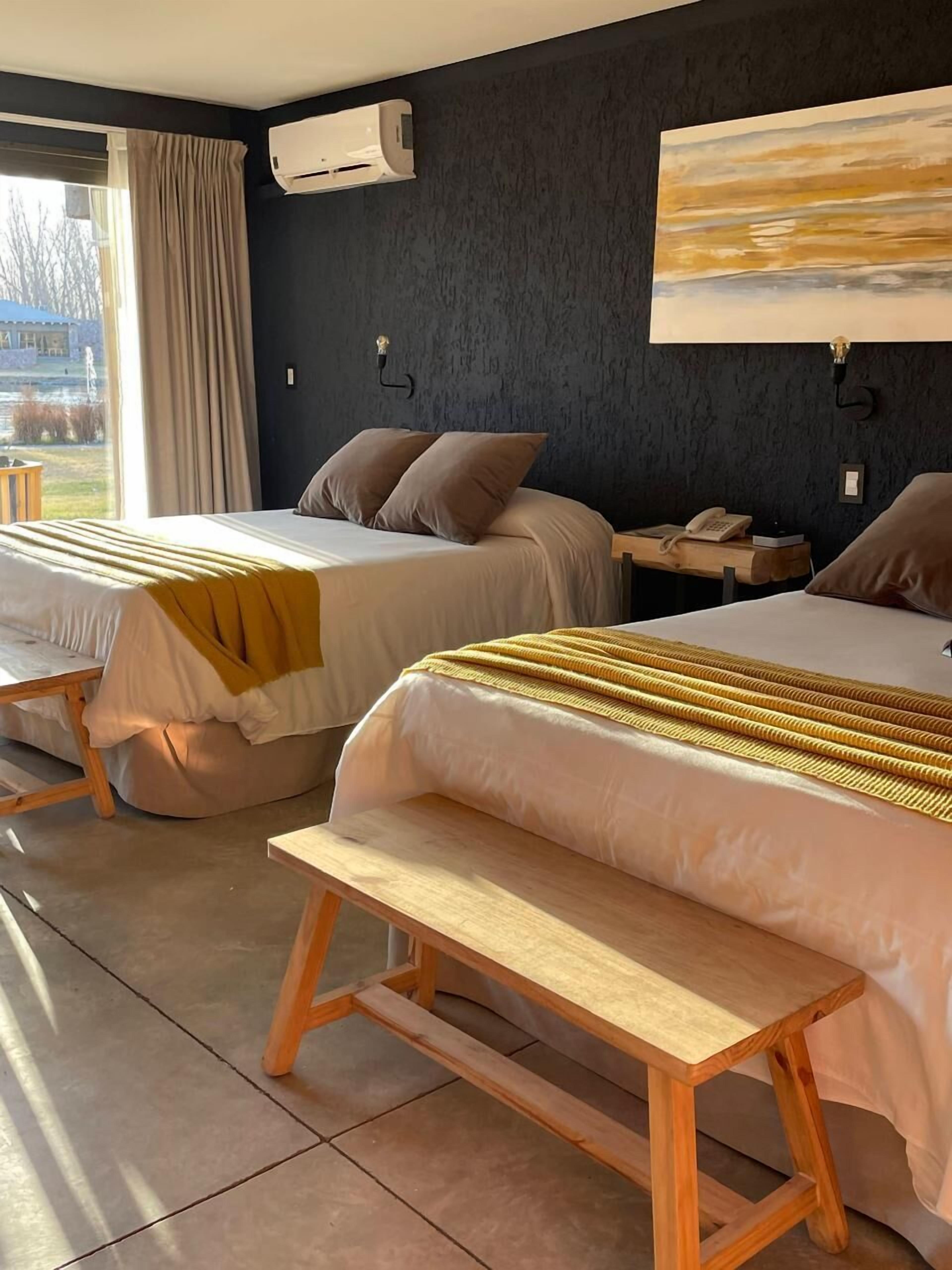 A hotel room with two beds, a bench, a bedside table with a lamp, and an air conditioning unit on the wall.