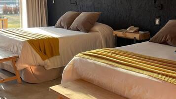 A hotel room with two beds, a bench, a bedside table with a lamp, and an air conditioning unit on the wall.