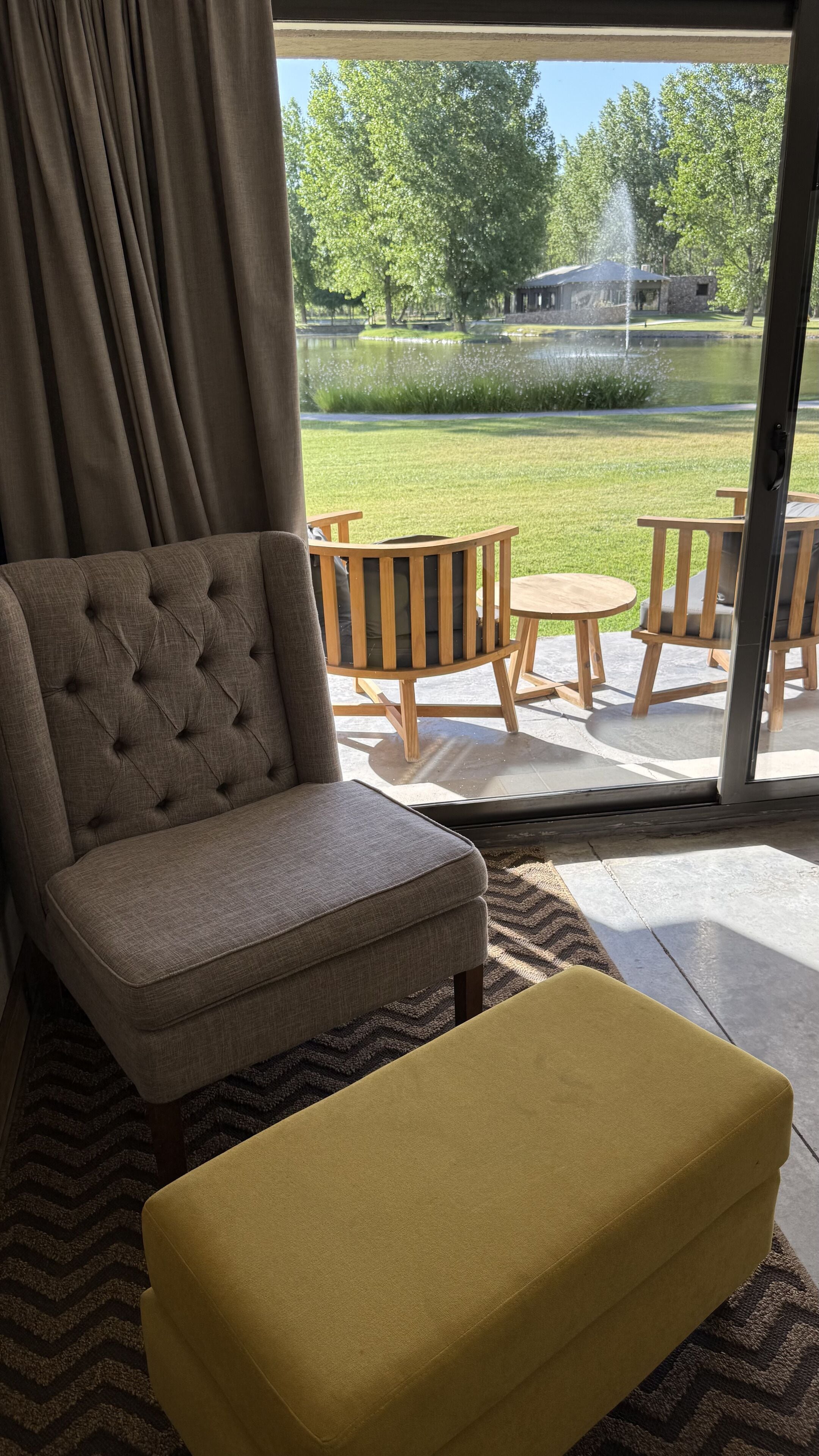 Ein Zimmer mit einem gepolsterten Sessel, einem gelben Hocker und Blick auf eine Terrasse mit Holzmöbeln und einem Teich.