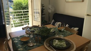 Un coin repas avec une table dressée pour deux personnes, agrémenté d’une nappe à motifs tropicaux, d’un ventilateur et offrant une vue sur un balcon verdoyant.