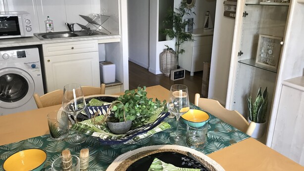 Un coin repas avec une table dressée pour deux personnes, un lave-linge et un espace cuisine équipé de placards et d’appareils électroménagers.
