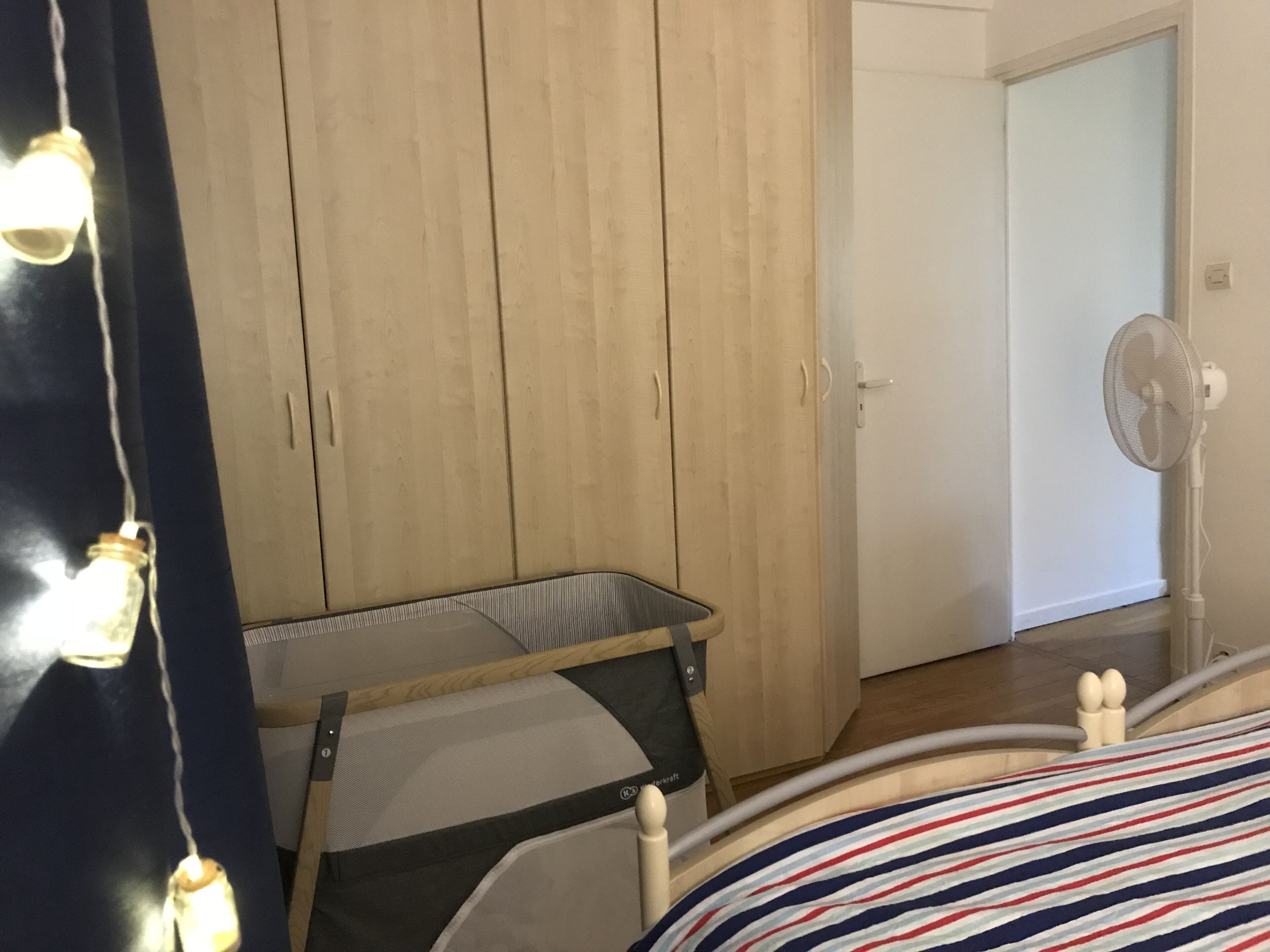 Een slaapkamer met een bed, een houten garderobe, een tafeltje en een staande ventilator.