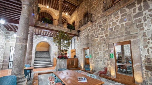 A stone-walled interior with a wooden table and chairs, a staircase, and a balcony with plants. - OYO Hostal Mich Morelia (Morelia)