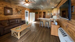 A cozy cabin interior with a wooden floor, a brown leather sofa, a wooden coffee table, a kitchen area with a refrigerator, and a ceiling fan. - AMAZING VIEW of Mount Magazine! (Booneville)