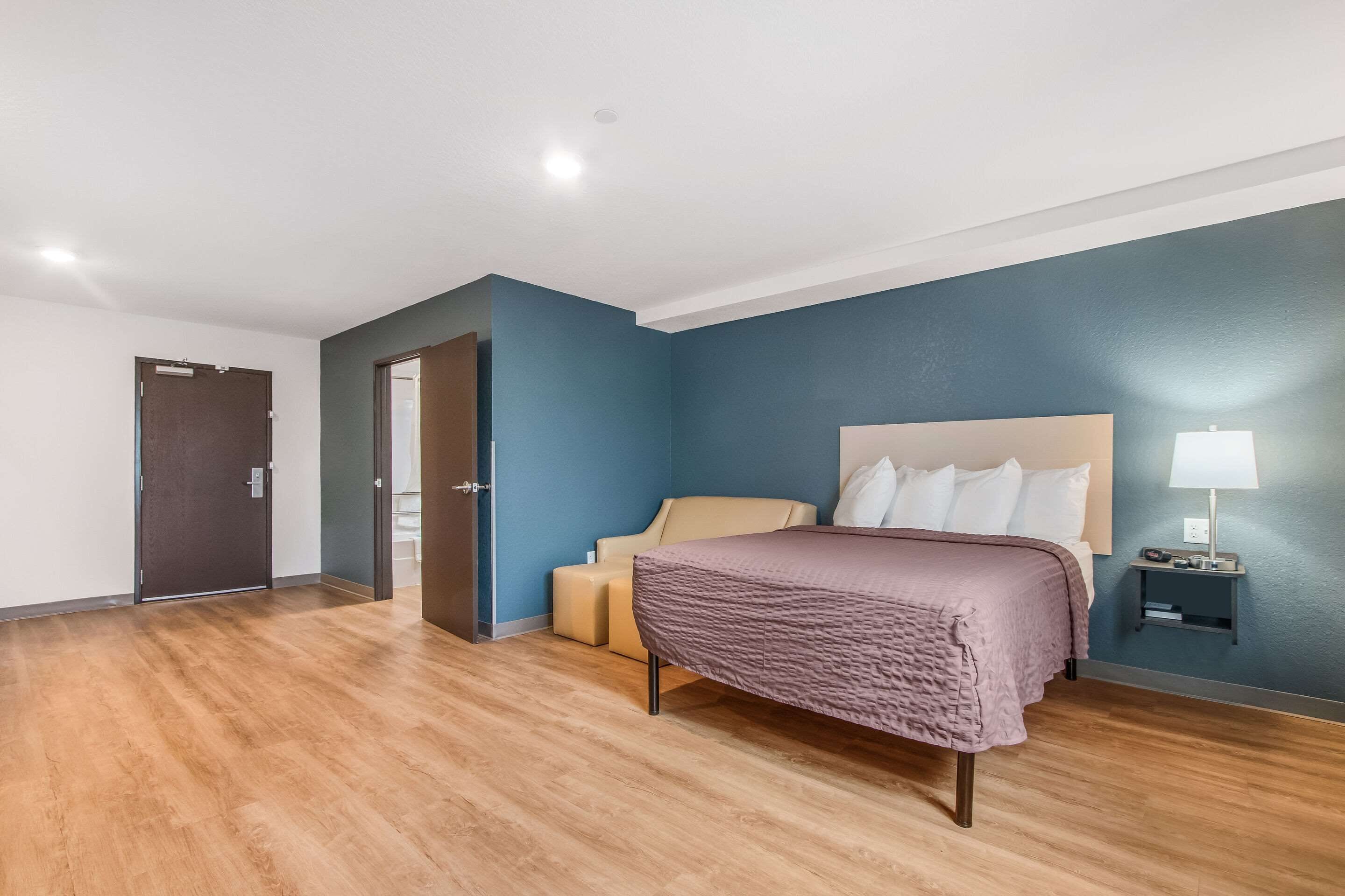 Una camera d'albergo moderna con un letto grande, un comodino con una lampada e una porta che dà su un'altra stanza.