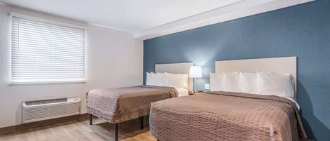 A hotel room with two beds, a window with blinds, a wall heater, and a blue accent wall.