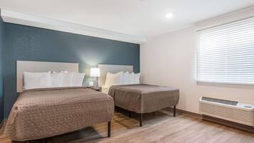 Quarto de hotel com duas camas, uma mesinha de cabeceira com abajur, uma janela com persianas e um ar-condicionado fixado na parede.