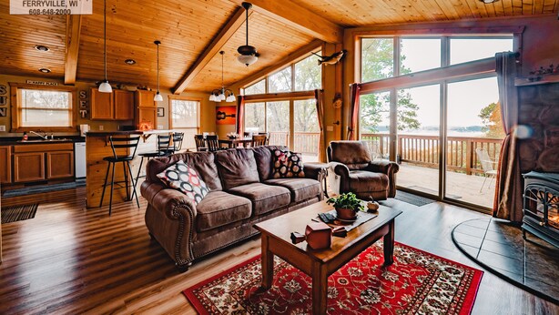 Ein gemütliches Blockhaus-Interieur mit Holzbalkendecke, einem Essbereich, einem Wohnzimmer mit Sofa und Sesseln sowie einem großen Fenster mit Blick auf eine Terrasse.