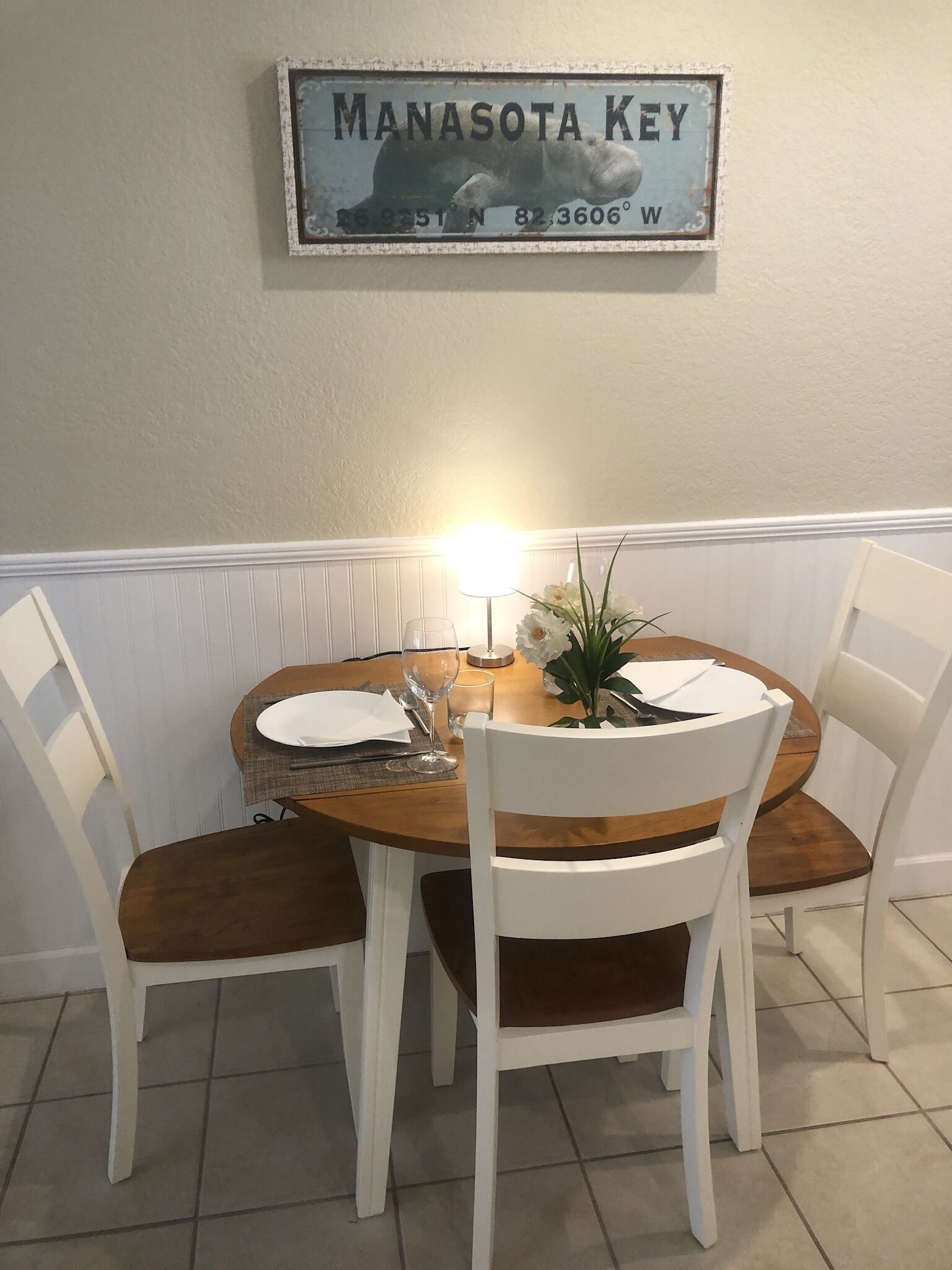 A dining area with a round wooden table, white chairs, and a wall-mounted sign reading "MANASOTA KEY".