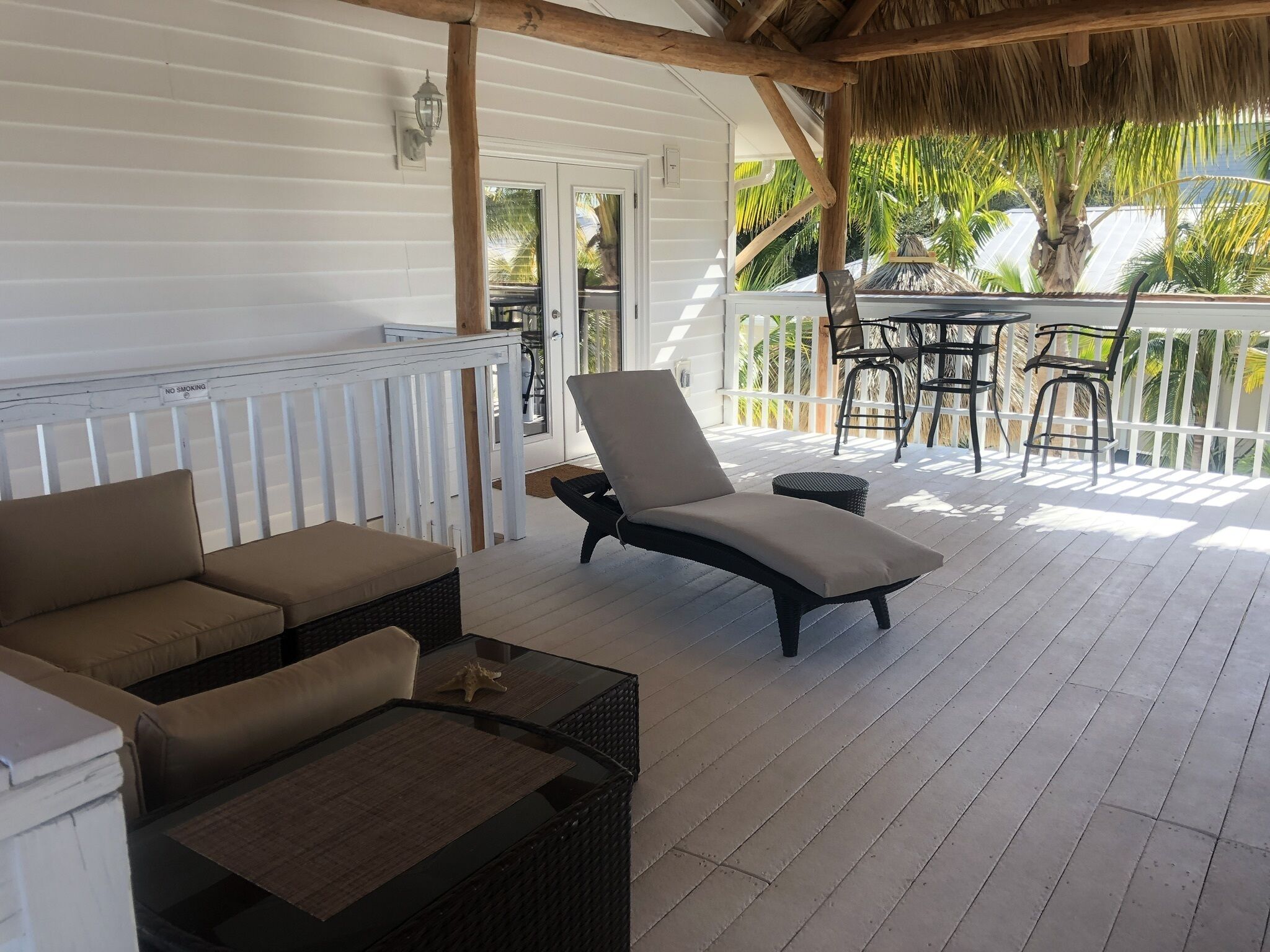 A covered outdoor seating area with a white railing, a lounge chair, a small table, and a dining table with chairs.