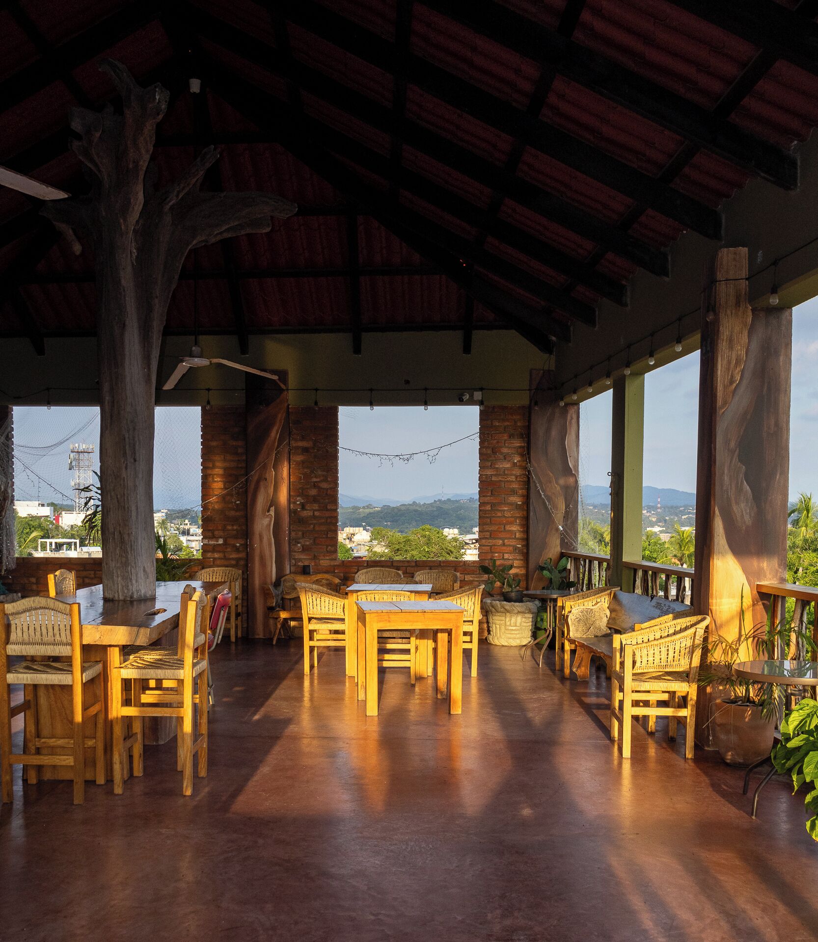 Un área de comedor al aire libre con mesas y sillas de madera, un tronco de árbol grande como centro de mesa, y vistas del paisaje.