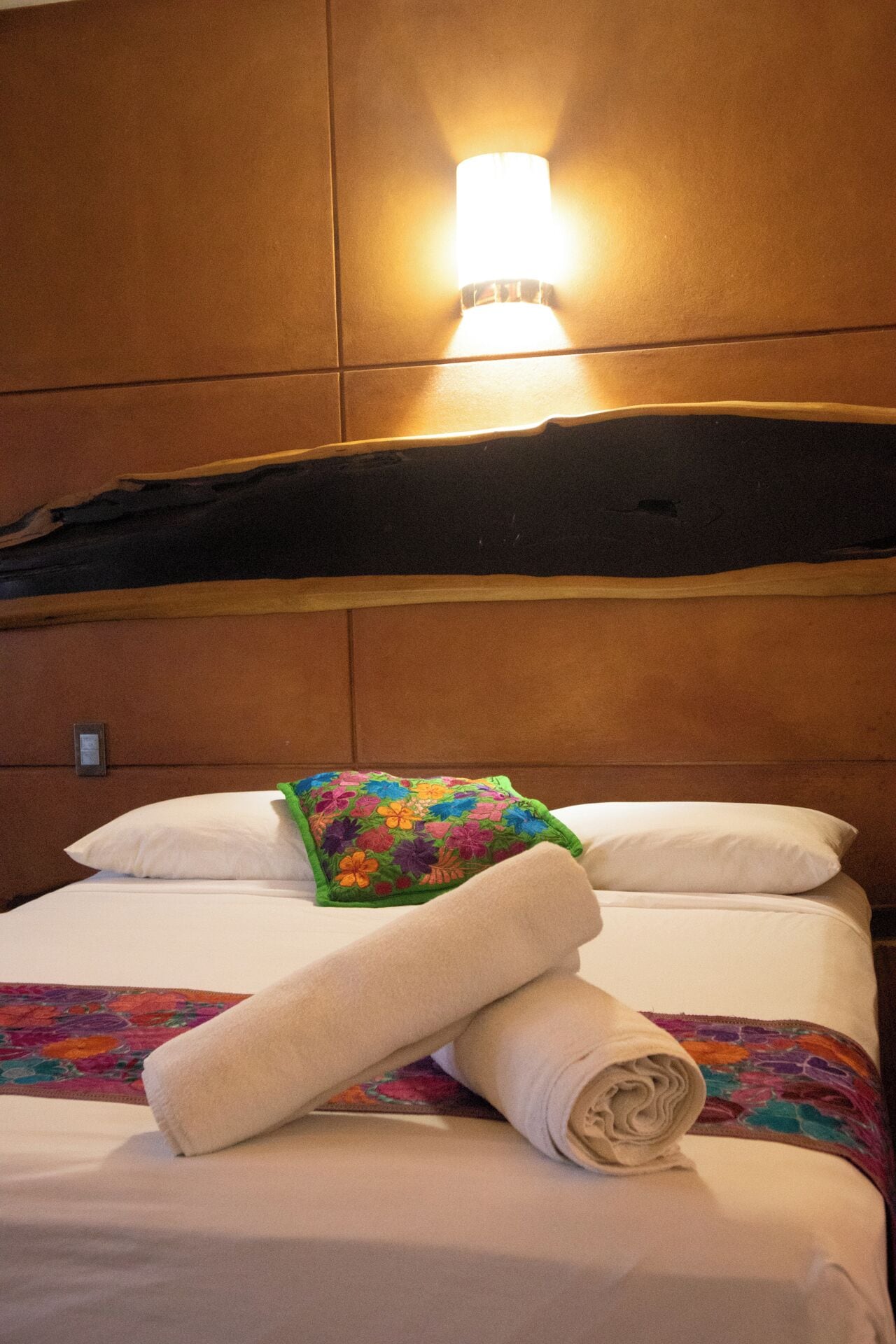 A hotel room with a bed, pillows, a colorful floral pillow, a rolled towel, and a wall-mounted light fixture.