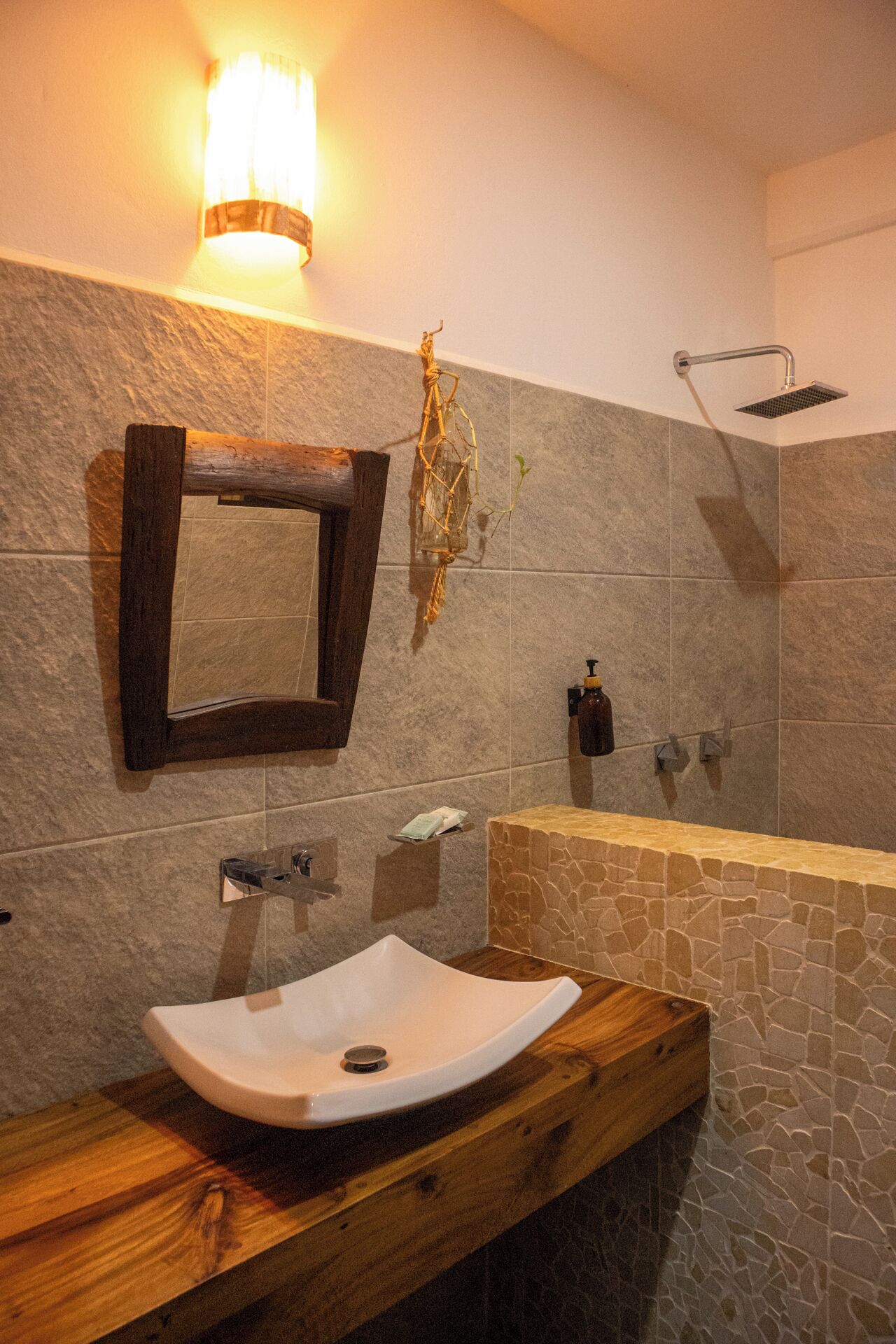 Un baño con un lavabo de piedra, un tocador de madera, un espejo, un cabezal de ducha y una lámpara de pared.