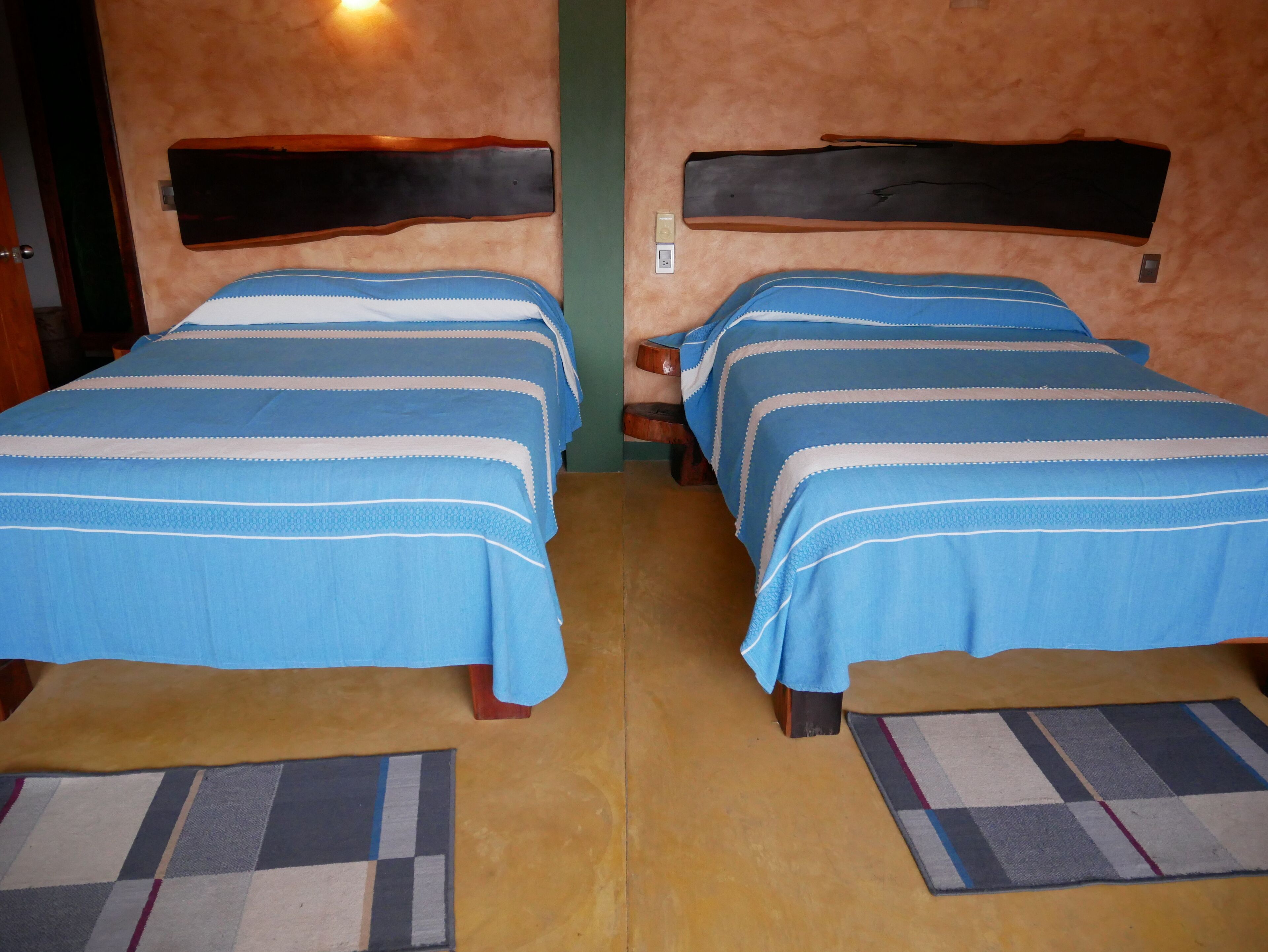 Dos camas individuales con ropa de cama azul y cabeceros de madera, una alfombra a cuadros y suelo de madera.