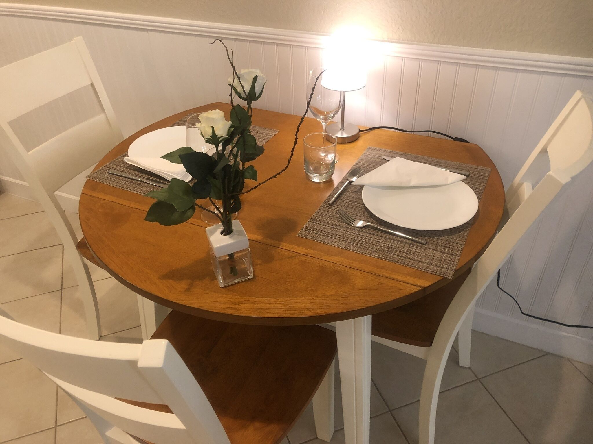 Una mesa de comedor redonda de madera con sillas blancas, un florero con una planta y una vela encendida.