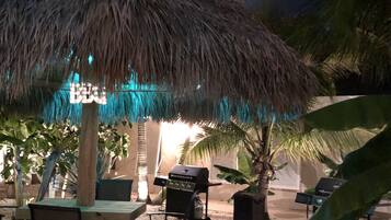 A thatched-roof outdoor seating area with tables and chairs, a grill, and a sign that reads "BBQ".