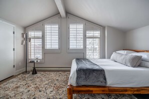 Un dormitorio con techo inclinado, una cama con ropa de cama blanca, un tapete estampado y una mesita de noche con lámpara.