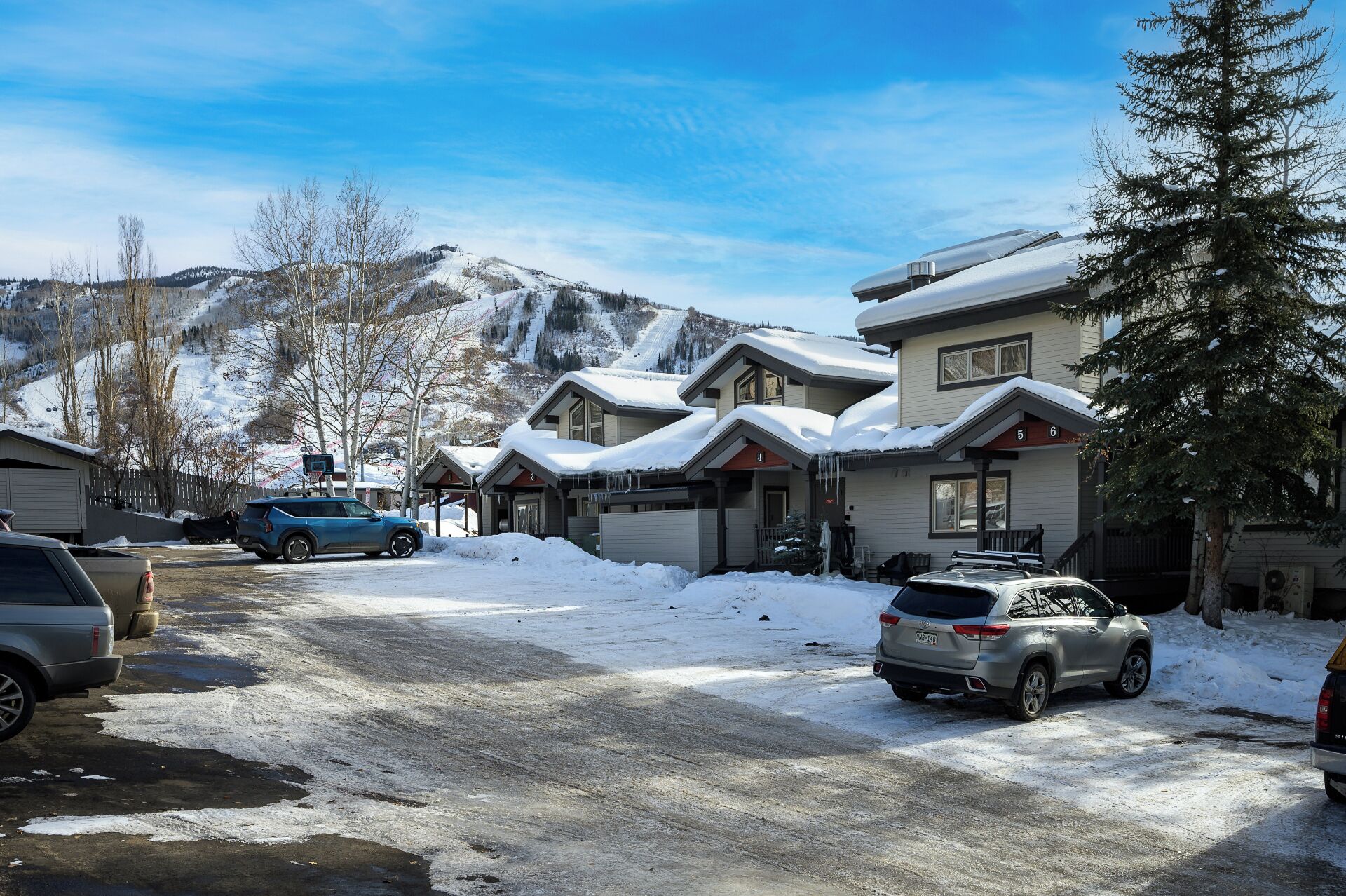A row of two-story buildings with snow-covered roofs, parked cars, and a mountainous backdrop.