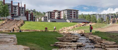 Un hôtel moderne avec un sentier de pierre menant à une petite cascade, entouré d’une végétation luxuriante et sous un ciel dégagé.