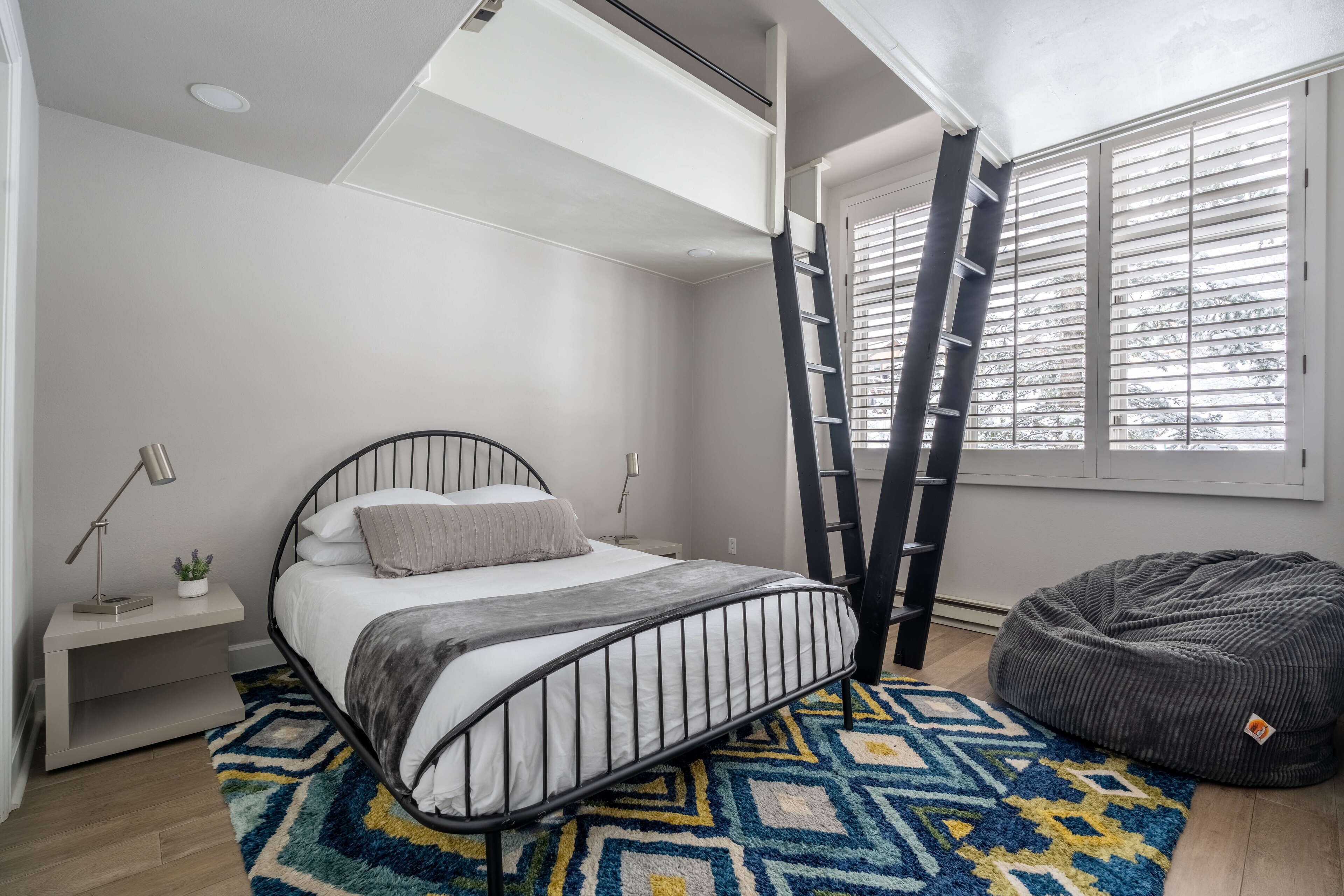 A bedroom with a bed, a bedside table, a lamp, a window with blinds, a ladder, and a bean bag chair.