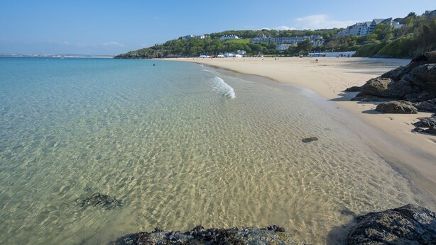 A beach with clear water, sandy shore, and buildings on a hill in the background.