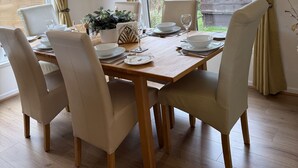 A dining area with a wooden table set for six, surrounded by six chairs, with a large window providing a view of greenery outside. - Cheviot Luxury Lodge (Morpeth)