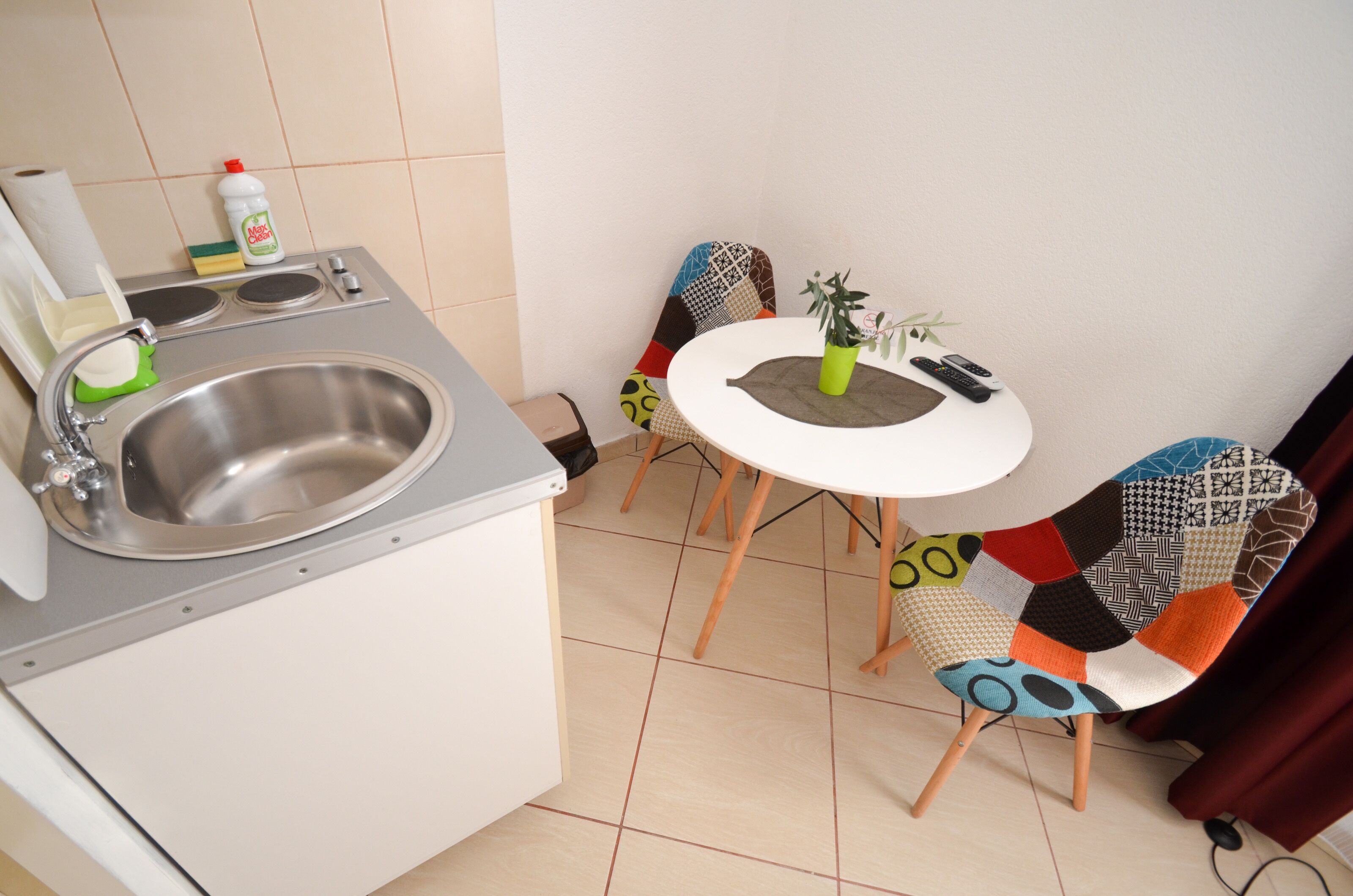 A small kitchenette with a stainless steel sink, a white countertop, and a small table with a potted plant. There are two chairs with colorful patterns.