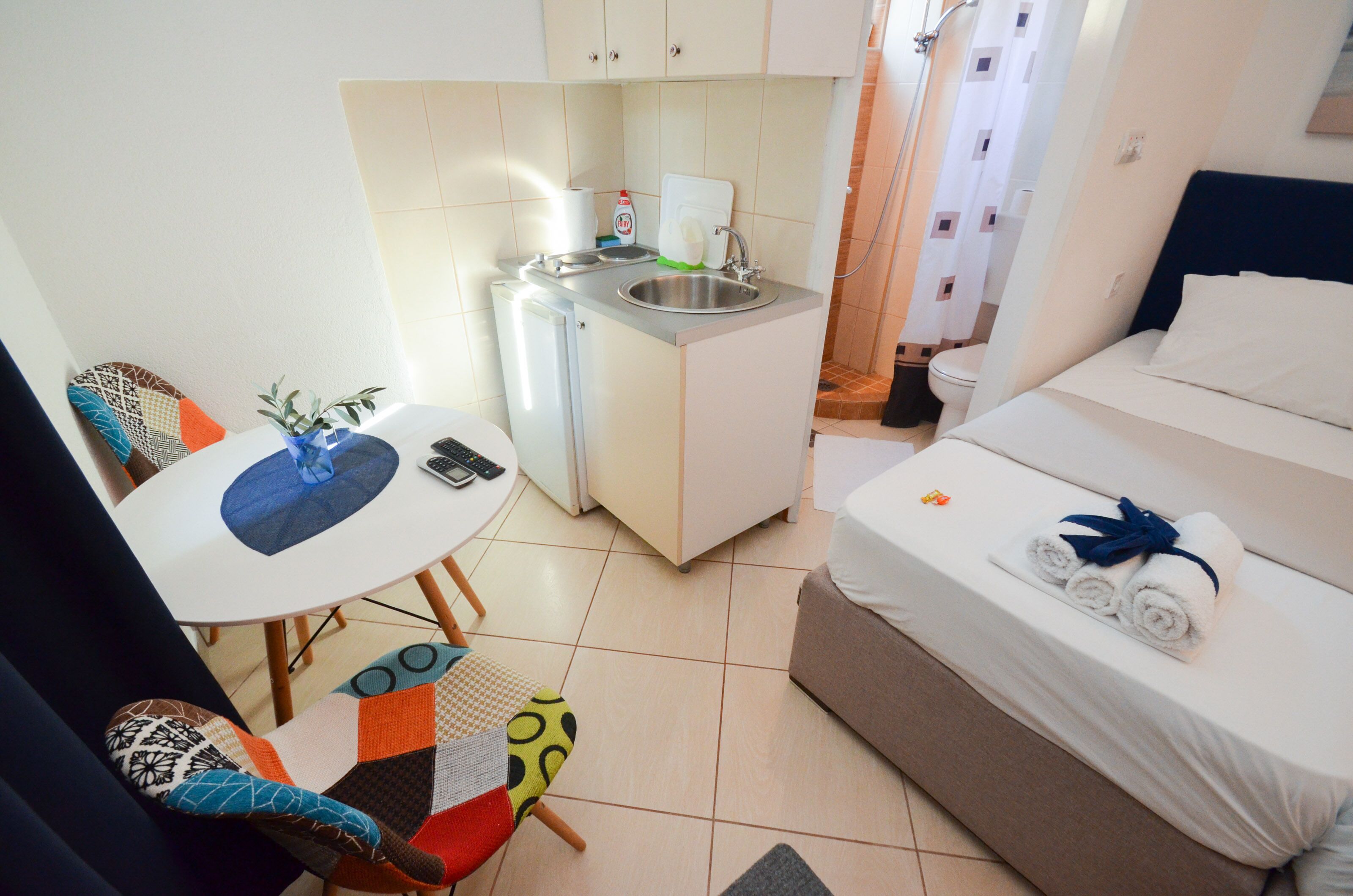 Habitación de hotel compacta con kitchenette, una cama individual con toallas, una pequeña mesa con sillas y zona de ducha.