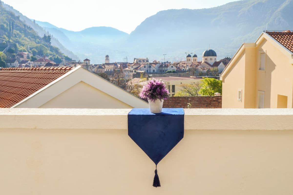 Ein Balkon mit einer blauen Tischdecke und einem Blumenstrauß, mit Blick auf eine Stadt mit historischen Gebäuden und Bergen im Hintergrund.