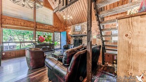 A cabin interior with a stone fireplace, leather seating, wooden beams, and large windows offering a view of the outdoors.
