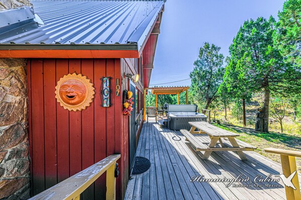 Una terraza de madera con mesas de picnic, un muro de piedra y un edificio rojo con un adorno de sol.