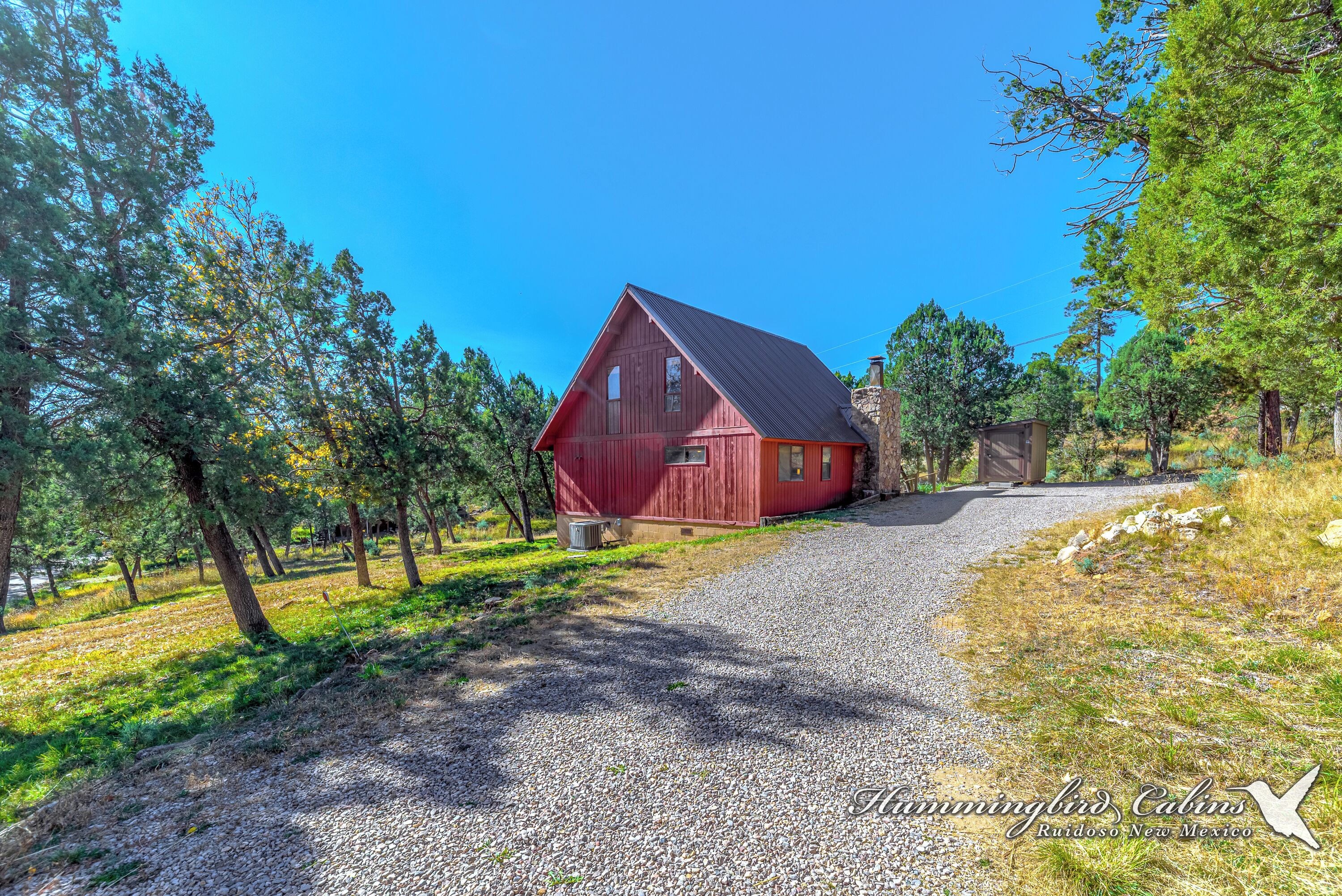 A red barn with a gravel driveway leading to it, surrounded by trees and a clear blue sky.