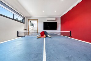 Une pièce avec une table de ping-pong, un téléviseur fixé au mur et une fenêtre donnant sur un paysage verdoyant.