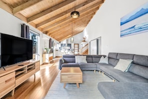 A spacious living room with a large flat-screen TV, wooden flooring, a grey sectional sofa, and a wooden coffee table.