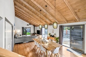 Un espace de vie moderne avec un plafond en bois, un coin repas comprenant une table et des chaises en bois, un canapé gris et un téléviseur en arrière-plan.